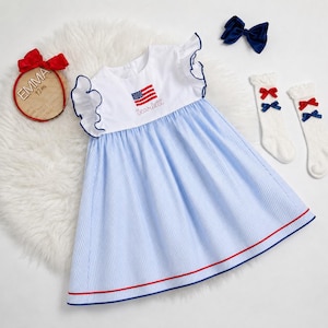 Könnte beinhalten: Ein patriotisches Babykleid mit einem weißen Oberteil mit einer gestickten amerikanischen Flagge und dem Namen "Scarlett". Der blau-weiß gestreifte Rock ist rot und blau eingefasst. Zubehör umfasst eine marineblaue Schleife und weiße Socken mit roten und blauen Schleifen.