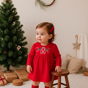 Può includere: Una bambina con un vestito rosso con una iniziale ricamata bianca e un motivo floreale. L'abito ha maniche lunghe e colletto Peter Pan. La bambina indossa calzini rossi e scarpe bianche. Un albero di Natale e regali confezionati sono sullo sfondo.