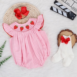 May include: Pink and white gingham baby onesie with embroidered strawberries. The onesie has short sleeves and a ruffled neckline. A red bow is on a woven basket in the background.