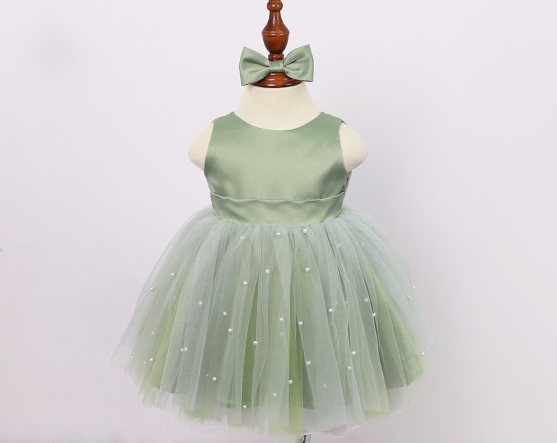 May include: A sage green tulle dress with a satin bodice and pearl embellishments. The dress has a bow on the top.