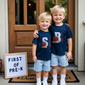 May include: Two children in navy blue t-shirts and blue gingham shorts. The shirts have a large letter, a red apple graphic, and the child's name. A sign reads "FIRST OF PRE-K".