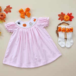 May include: A pink smocked dress with a Halloween-themed embroidered neckline featuring a pumpkin, ghost, spider, and candy corn. The dress has short puffed sleeves and a gathered skirt. The dress is paired with white knee-high socks with orange bows.