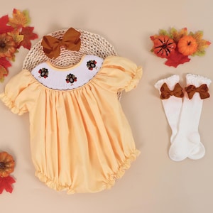 May include: A yellow baby smocked dress with a turkey embroidery design. The dress has puffed sleeves and a gathered skirt. The dress is paired with a brown bow headband and white knee-high socks with brown bows.