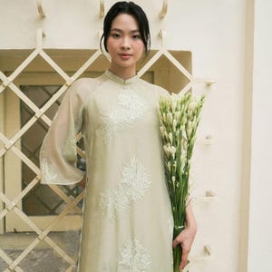 Puede incluir: Un vestido verde claro con bordado floral blanco y cuello mandarín. El vestido tiene mangas largas y transparentes y se combina con un ramo de flores blancas. El vestido es de estilo tradicional, adecuado para ocasiones especiales.