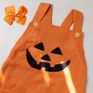 May include: Orange knitted baby overalls with a black pumpkin face design. The overalls have two straps and a button closure.