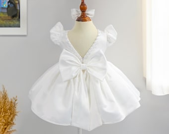 May include: White satin baby dress with a large bow on the back and a pearl trim around the neckline. The dress has short puffed sleeves.