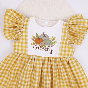 May include: A yellow and white gingham dress with frilled sleeves. The dress has a white bib with a pumpkin design and the name "Everly" in brown script. The dress is designed for a child.