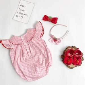 May include: A pink and white gingham baby romper with red trim, paired with a red bow, headband, and shoes, all adorned with strawberry accents. The image includes a sign that reads "buy less, choose well."