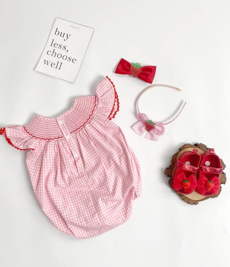 May include: A pink and white gingham baby romper with red trim, paired with a red bow, headband, and shoes, all adorned with strawberry accents. The image includes a sign that reads "buy less, choose well."