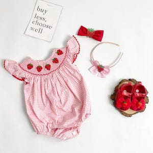 May include: A pink gingham baby romper with red strawberry embroidery, paired with a matching red bow, headband, and shoes. The shoes are adorned with strawberry accents. A card with the text "buy less, choose well" is also present.
