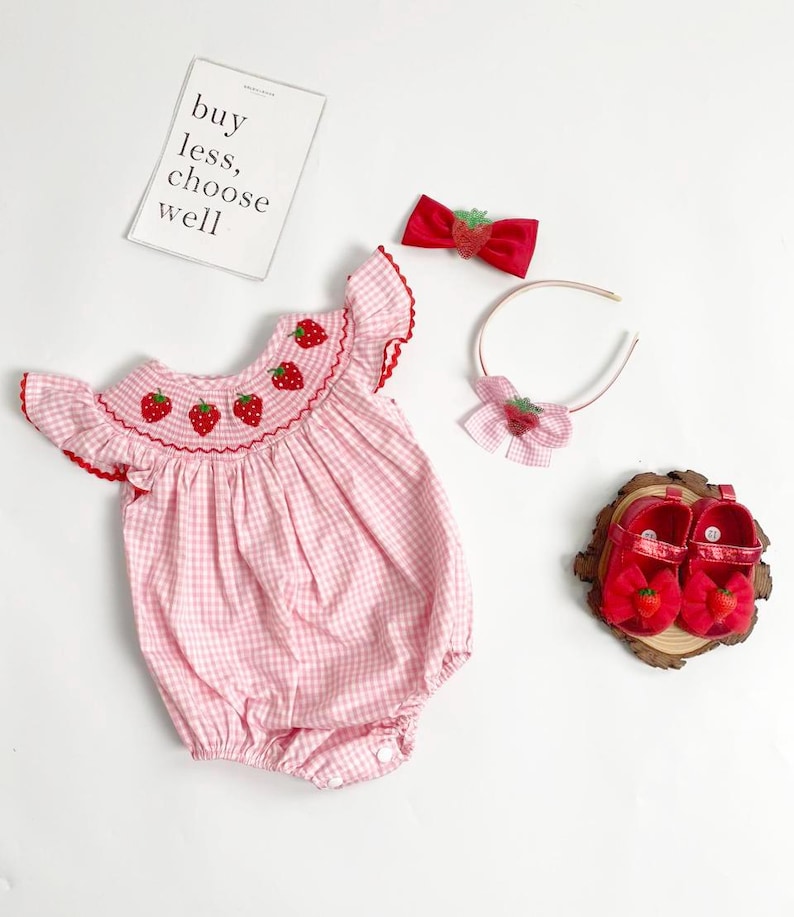May include: A pink gingham baby romper with red strawberry embroidery, paired with a matching red bow, headband, and shoes. The shoes are adorned with strawberry accents. A card with the text "buy less, choose well" is also present.