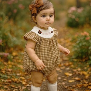 May include: A baby wearing a brown gingham romper with a white collar embroidered with the name "OCTAVIA". The outfit is paired with brown shoes, white socks, and a brown bow. The background is a blurred outdoor scene with fall foliage.