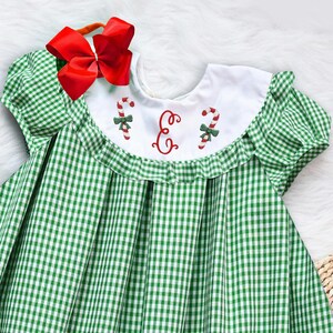 Può includere: Un vestito a quadri vichy verde e bianco con colletto bianco e maniche corte. Il colletto è ricamato con bastoncini di zucchero rosso e bianco e la lettera "E" ricamata al centro.