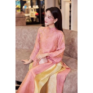 May include: A pink and yellow traditional dress with long sleeves and a high collar. The dress has a textured appearance and is worn by a person sitting on a sofa. The dress is a blend of pink and yellow hues.