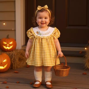 Könnte beinhalten: Ein kleines Kind in einem gelben Vichy-Kleid und passendem Stirnband, das einen kleinen Weidenkorb hält. Das Kleid hat einen weißen Kragen mit floraler Stickerei. Zwei geschnitzte Kürbisse und Heuballen schmücken die Veranda für Halloween.