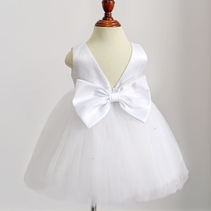 May include: White tulle dress with a large white satin bow on the back. The dress is decorated with small white beads.