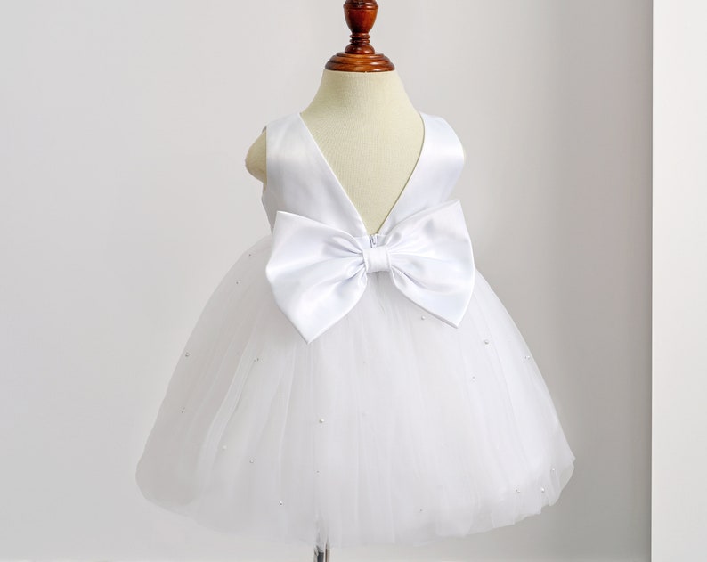 May include: White tulle dress with a large white satin bow on the back. The dress is decorated with small white beads.