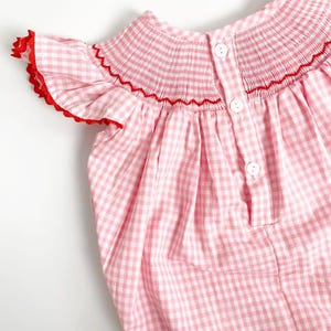 May include: A pink and white gingham baby dress with red trim. The dress features a smocked bodice with red stitching and small white buttons down the back. The sleeves have a ruffled edge with red trim.