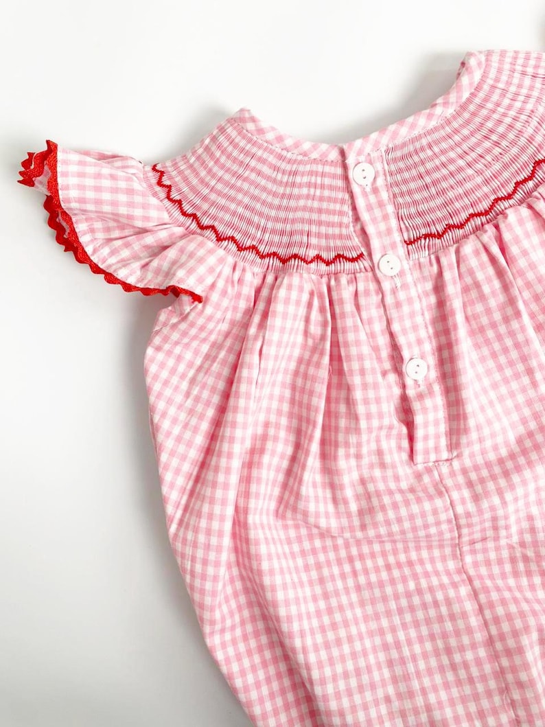 May include: A pink and white gingham baby dress with red trim. The dress features a smocked bodice with red stitching and small white buttons down the back. The sleeves have a ruffled edge with red trim.