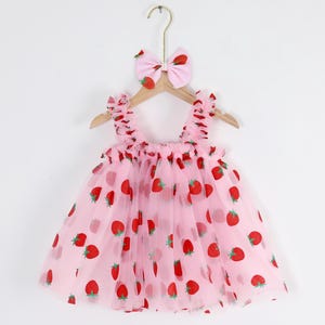 May include: A pink tulle dress with a strawberry print and matching pink bow headband. The dress has a sleeveless design and a ruffled neckline.