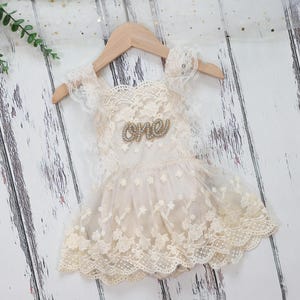 May include: A cream lace dress with a floral pattern, displayed on a wooden hanger. The dress has a 'one' embellishment and lace sleeves. The dress is designed for a baby's first birthday.