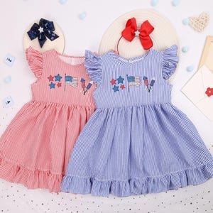 May include: Two sleeveless dresses with red and blue stripes. Each dress has a ruffled hem and flutter sleeves. Embroidered on the front are stars, flags, and fireworks. One dress is red, and the other is blue.