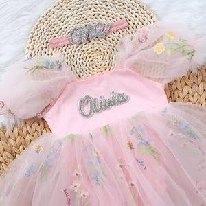 May include: Pink tulle dress with embroidered flowers and a silver glitter 'Olivia' name appliqué. The dress has puff sleeves and a matching headband with silver glitter 'one' appliqué.