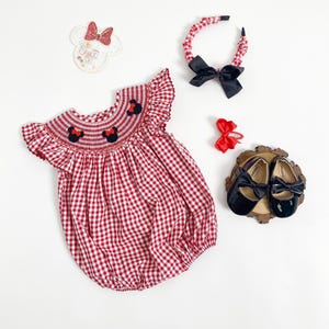 May include: A red and white gingham romper with Minnie Mouse embroidery, a matching headband with a black bow, and black patent leather shoes with bows. A red bow hair clip and a Minnie Mouse-shaped badge complete the set.