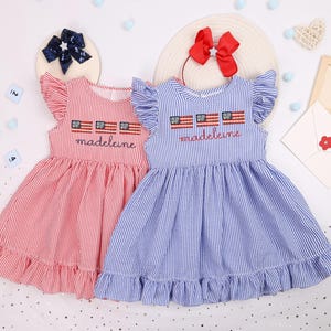 Puede incluir: Dos vestidos a rayas, uno rojo y otro azul, con mangas y dobladillos con volantes. Cada vestido tiene el nombre "Madeleine" bordado debajo de tres pequeñas banderas americanas. Los vestidos son sin mangas y tienen cuello redondo.