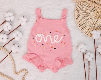 Puede incluir: Mono de bebé de punto rosa con un "uno" bordado en blanco y espolvoreado de colores.