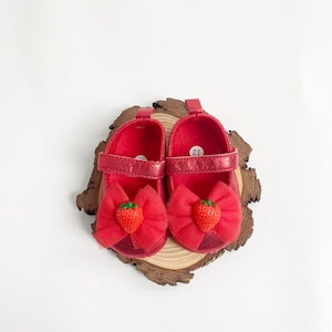 May include: A pair of red baby shoes with a bow and a strawberry accent. The shoes are size 12 and have a strap closure. The shoes are on a wooden slice.