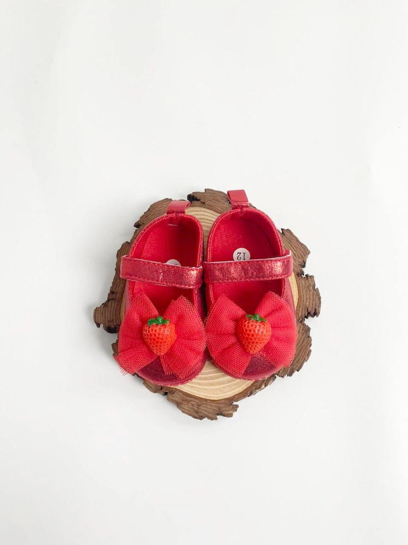 May include: A pair of red baby shoes with a bow and a strawberry accent. The shoes are size 12 and have a strap closure. The shoes are on a wooden slice.