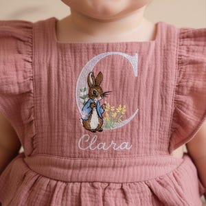 以下が含まれることがあります： ダスティローズ色のベビーロンパース。ピーターラビットのデザインが刺繍されています。 ロンパースは、フリルネックラインとストラップが特徴で、デザインの下に「Clara」という名前が刺繍されています。 デザインにはウサギ、花、文字「C」が含まれています。