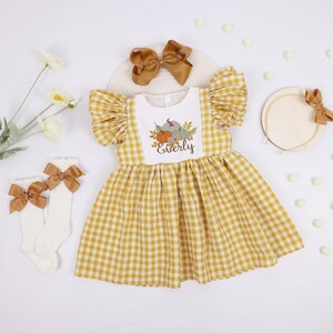 May include: A yellow and white gingham dress with short sleeves and a pumpkin graphic. The dress is paired with white socks with brown bows, a brown bow headband, and a matching hair bow. The word "Esterly" is printed on the dress.