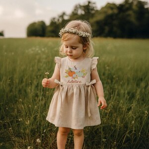May include: A beige linen dress featuring a pumpkin and leaf design, with the name "Adeline" embroidered. The dress has ruffled sleeves and tie details. A young child wearing the dress and a floral crown stands in a field of tall grass.
