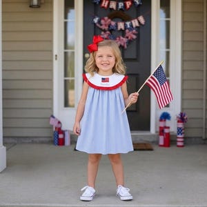 Könnte beinhalten: Ein junges Mädchen in einem blau-weiß gestreiften Kleid mit rotem und weißem Kragen, das eine kleine amerikanische Flagge hält. Sie hat eine rote Schleife im Haar. Das Kleid hat ein kleines amerikanisches Flaggenemblem am Kragen.