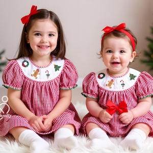 Op de afbeelding: Twee jonge kinderen in bijpassende rood-witte geruite kerstjurken met geborduurde kragen. De jurken hebben peperkoekmannetjes en zuurstokontwerpen. Elk kind draagt een rode strik in het haar.