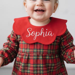 Può includere: Un vestito a quadri rossi con un colletto rosso abbinato ricamato con il nome "Sophia" in corsivo bianco. L'abito ha maniche lunghe e un corpetto aderente. Il bambino sorride.