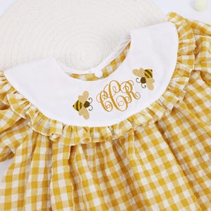 Könnte beinhalten: Gelb-weiß kariertes Babykleid mit weißem Kragen. Der Kragen ist mit gestickten Bienen und einem Monogramm verziert. Das Kleid hat Puffärmel und ein passendes Karomuster.