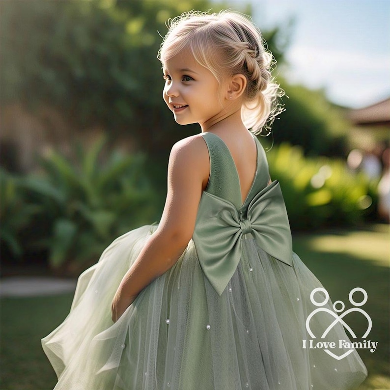 May include: A young girl wearing a sage green tulle dress with a large satin bow on the back. The dress is decorated with small pearl-like embellishments. The girl is smiling and looking to the side. The image has the text "I Love Family" at the bottom.