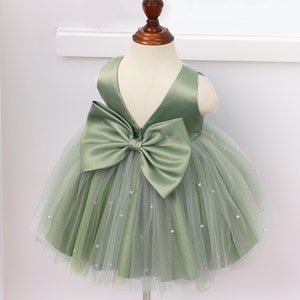 May include: A green tulle and satin dress with a large bow in the back. The dress is decorated with small white pearls.