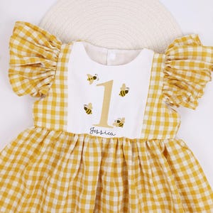 Puede incluir: Un vestido de cuadros amarillos y blancos con mangas con volantes. El vestido presenta un babero blanco con el número 1, abejas bordadas y el nombre "Jessica". El vestido está diseñado para el primer cumpleaños de un niño.