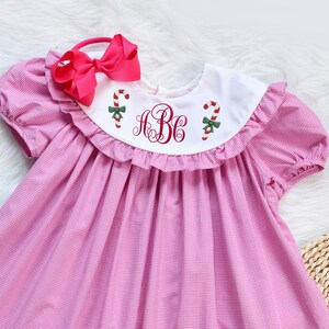 Può includere: Abito a quadri vichy rosa e bianco con colletto bianco Peter Pan e scollatura a balze. L'abito ha maniche corte ed è ricamato con una canna da zucchero e il monogramma "HBC".