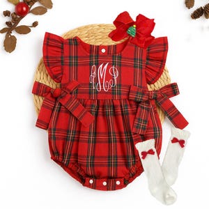 Könnte beinhalten: Rote Baby-Spielanzug mit Karomuster, weißem Monogramm und passenden Socken. Der Spielanzug hat eine Schleife auf dem Rücken und an den Ärmeln sowie eine große rote Schleife mit einem kleinen Weihnachtsbaum auf dem Kopf. Die Socken haben rote Schleifen.