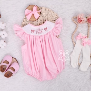 May include: Pink baby girl's smocked dress with a white collar and pink bows. The dress has a monogram "S.H." embroidered on the front. The dress is paired with a pink bow headband and white socks with pink bows.