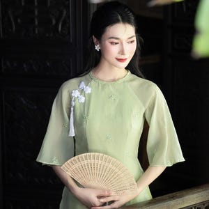 Può includere: Una donna indossa un abito verde chiaro con ricami floreali e maniche svasate, tenendo in mano un ventaglio beige intagliato in modo intricato. L'abito presenta uno scollo tondo e un abbellimento floreale bianco con nappa.