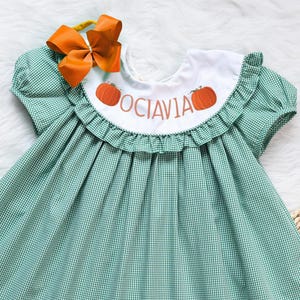 May include: A green and white gingham dress with a white Peter Pan collar and a ruffle trim. The dress has a pumpkin and the name "Octavia" embroidered on the collar.