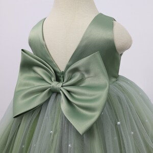 May include: A sage green tulle dress with a satin bow on the back. The dress is decorated with small pearl-like embellishments.