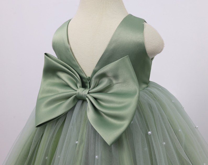 May include: A sage green tulle dress with a satin bow on the back. The dress is decorated with small pearl-like embellishments.
