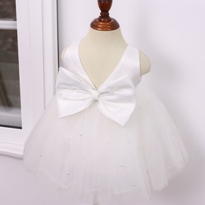 May include: White tulle baby dress with a large white satin bow on the bodice. The dress is decorated with small white beads.
